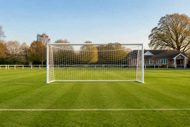 A white football goal on a sunlit green pitch with a clubhouse beyond